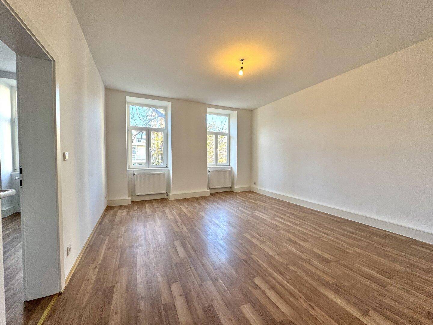 Geräumiges, unmöbliertes Wohnzimmer mit hellem Holzboden und zwei großen Fenstern.