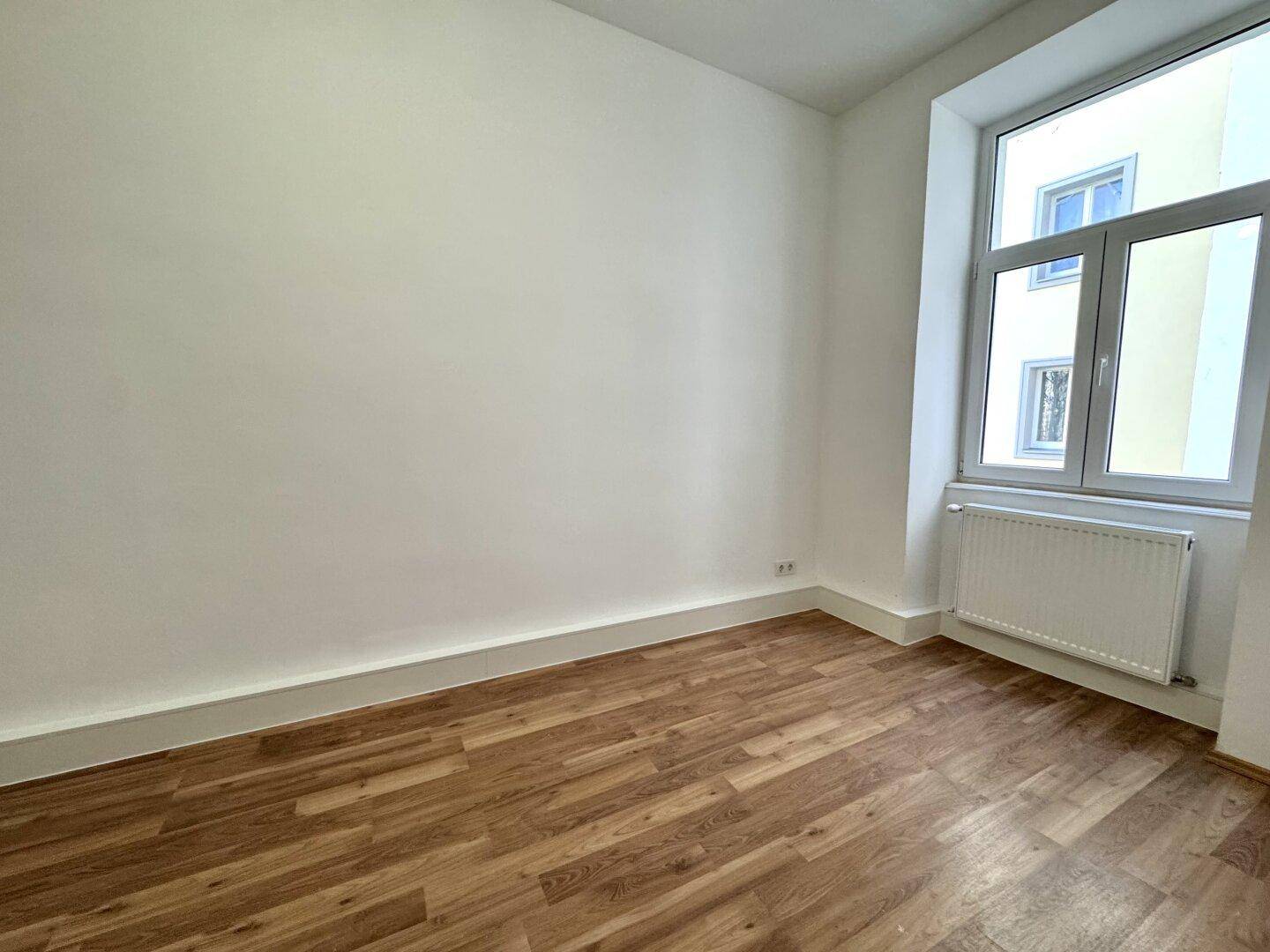 Leerer Raum mit Holzboden und einem großen Fenster, das viel Licht hereinlässt.
