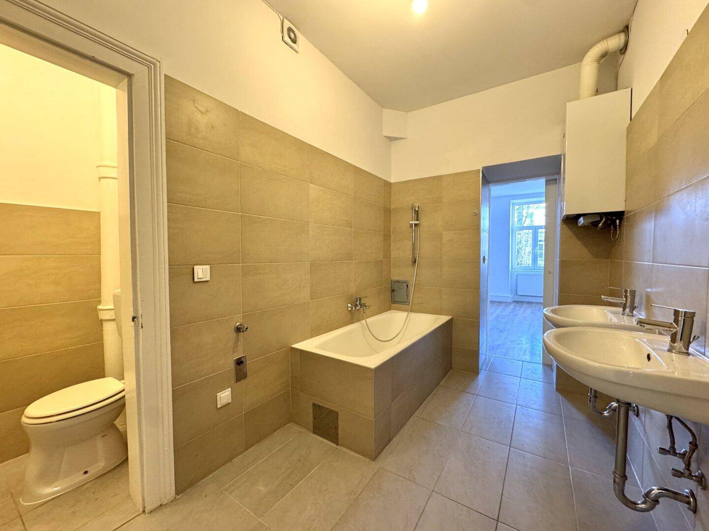 Badezimmer mit zwei Waschbecken, Badewanne und beigefarbenen Fliesen an Wänden und Boden.