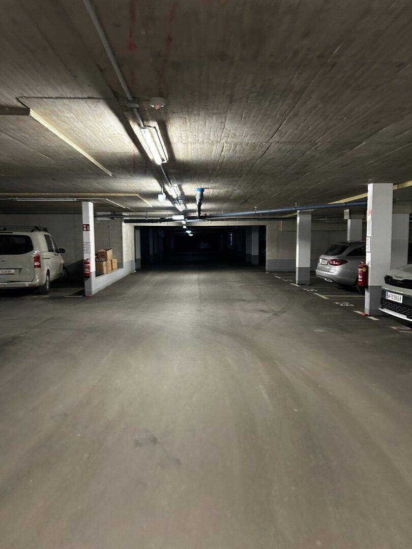 Langer Gang in einer Tiefgarage mit parkenden Autos und Lagerbereichen im hinteren Teil.