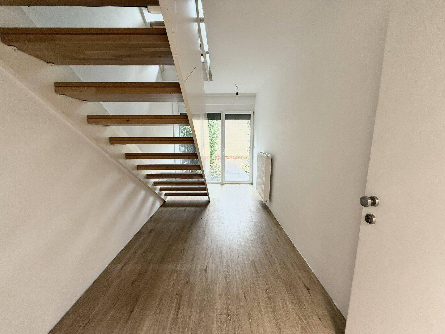 Heller Flur mit offener Holztreppe und Zugang zu einem Außenbereich durch eine Glastür.