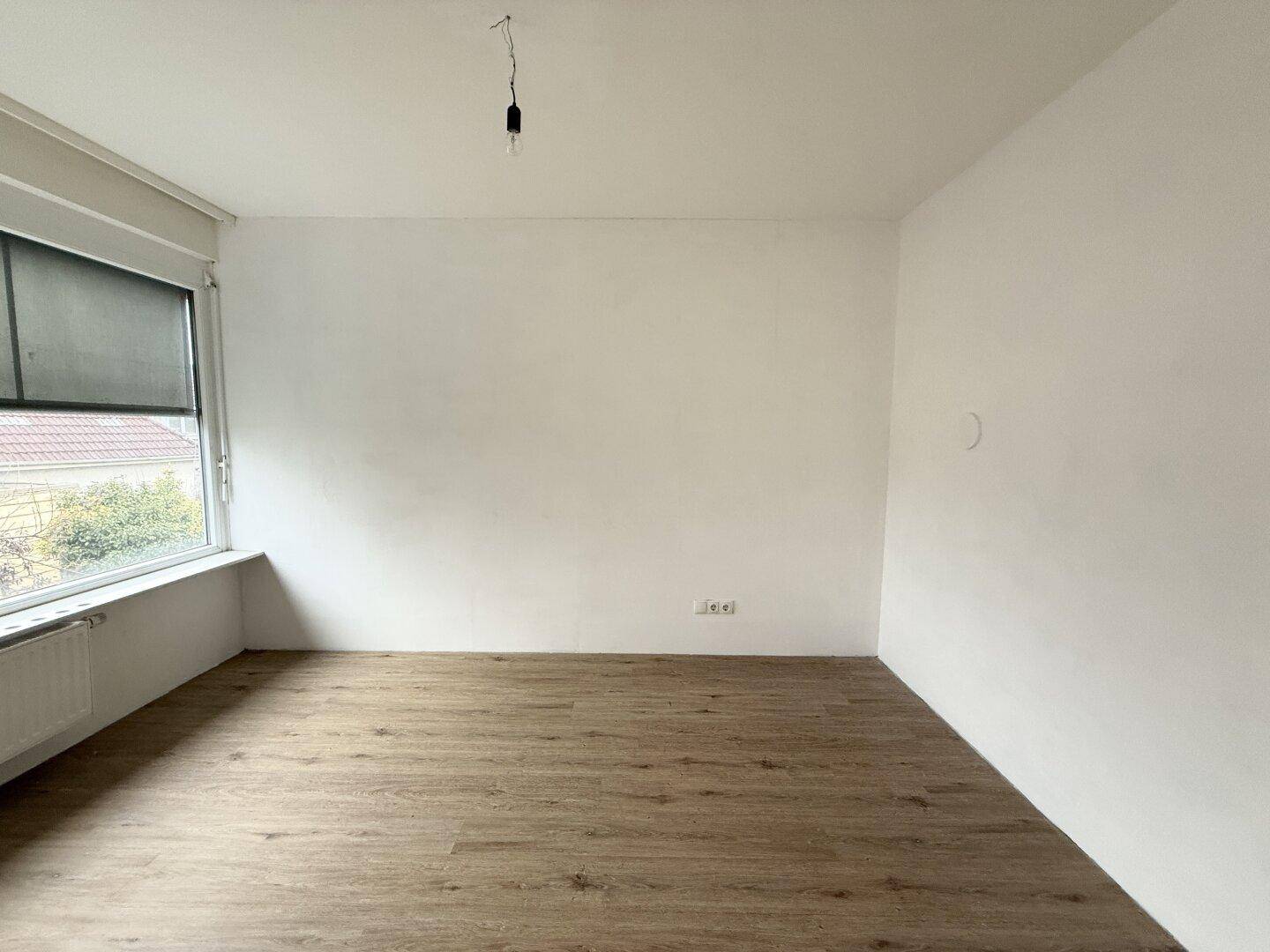 Helles Zimmer mit Laminatboden und großem Fenster, das viel Tageslicht hereinlässt und einen Ausblick bietet.
