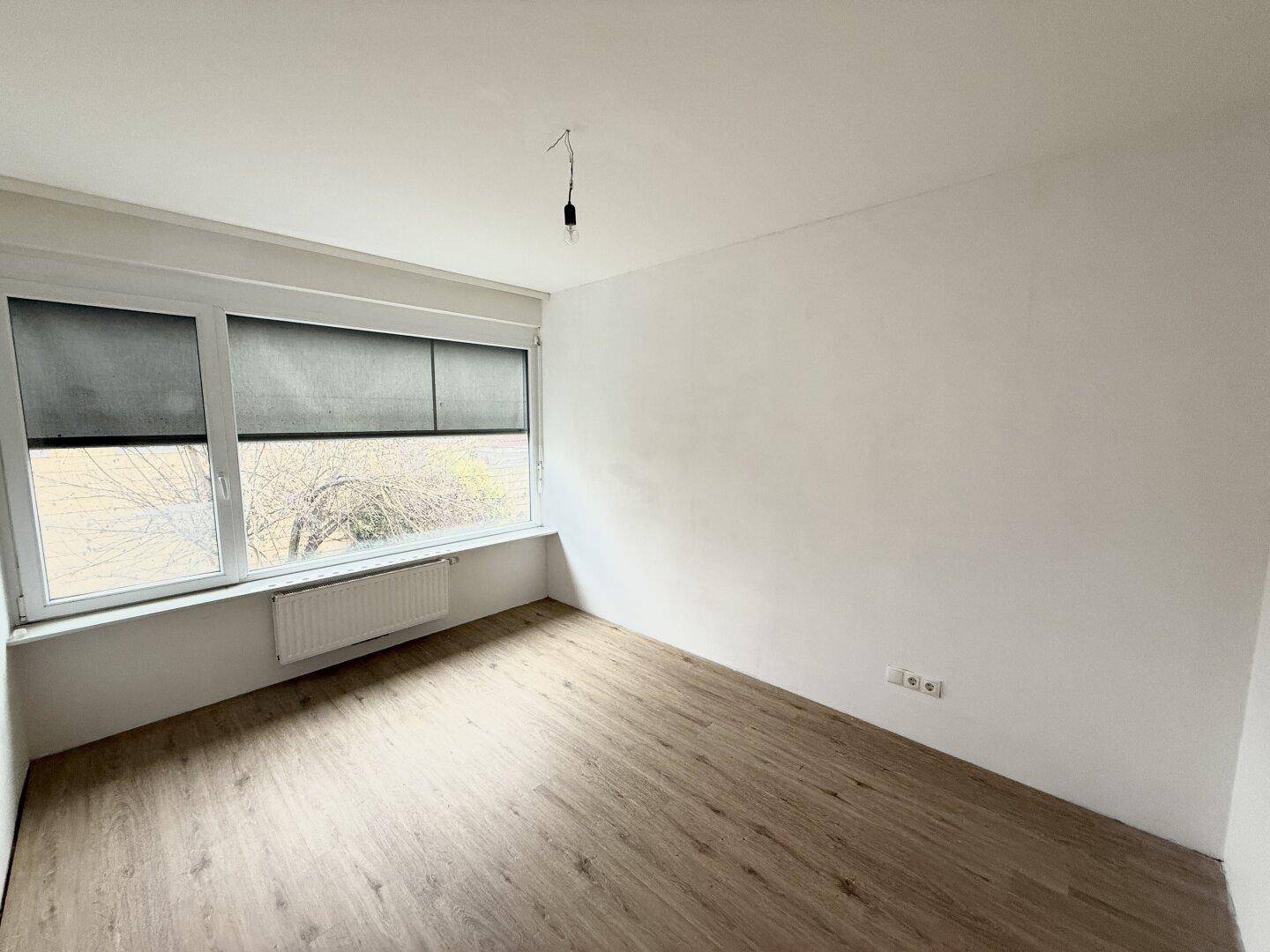 Freundliches Zimmer mit Laminatboden und breitem Fenster, ausgestattet mit einem Heizkörper unter der Fensterbank.