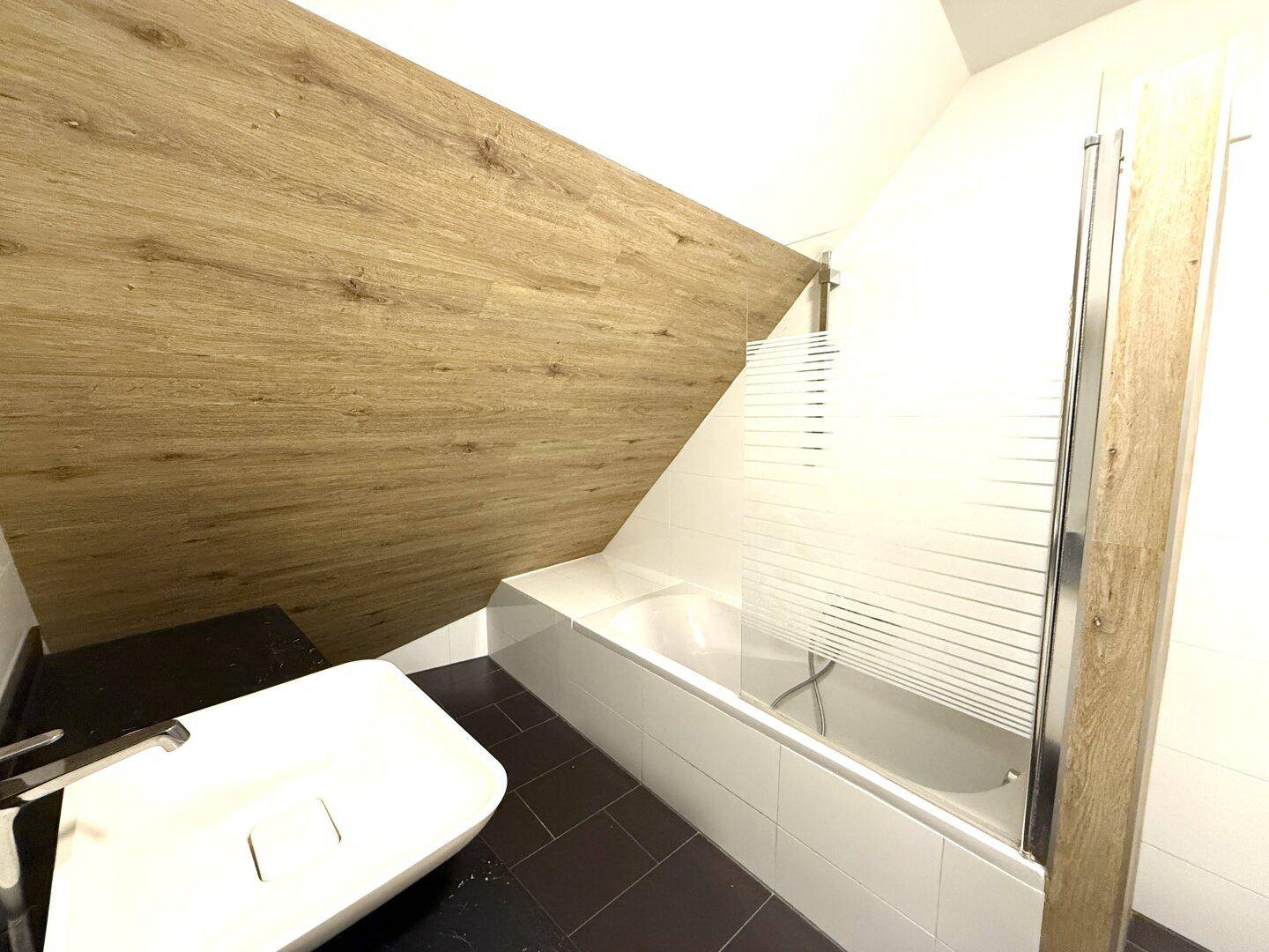 Badewanne mit Duschabtrennung in einem modernen Badezimmer unter der Dachschräge, mit Holzoptik-Wand.