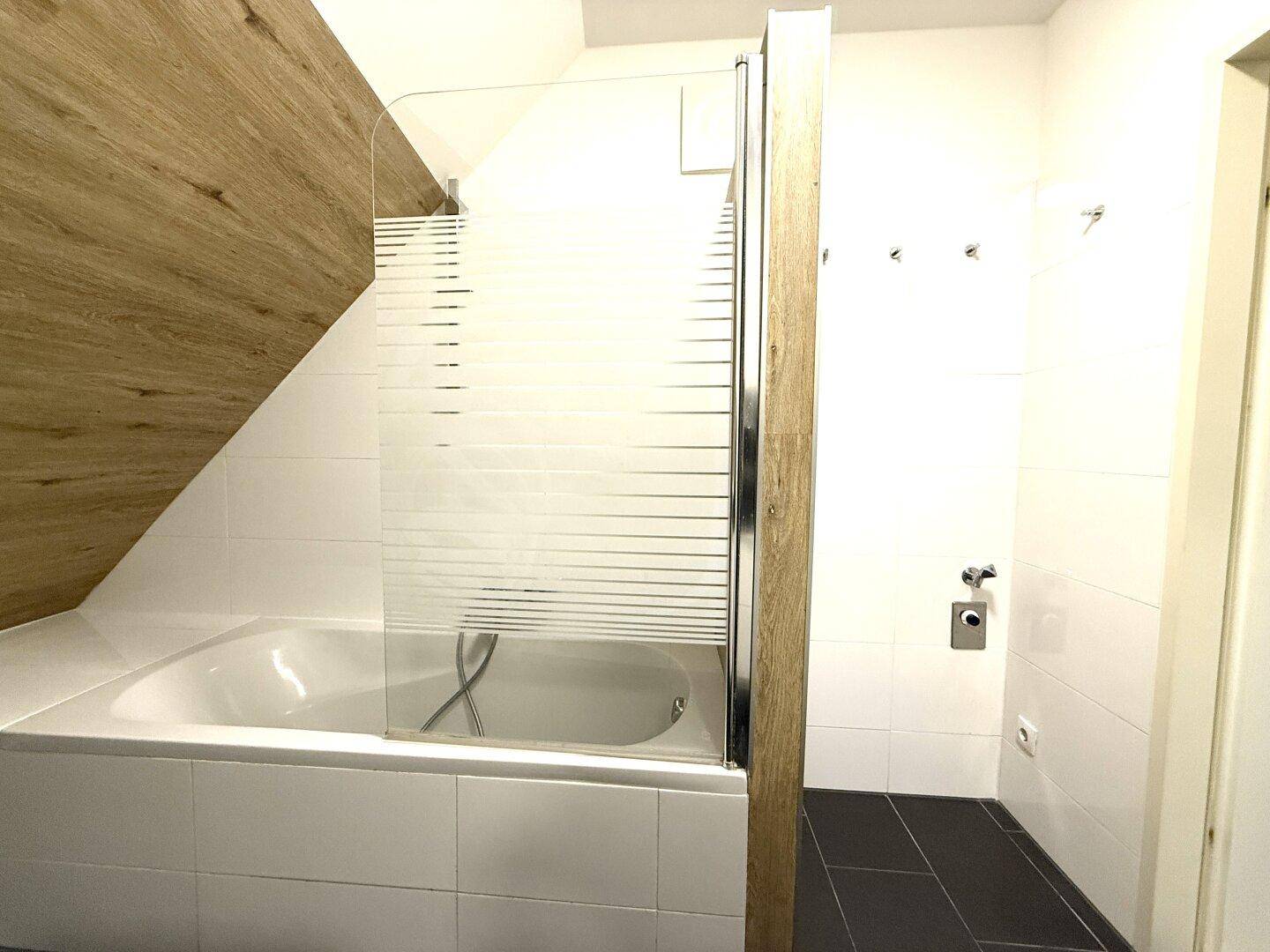 Badewanne mit Duschabtrennung in einem modernen Badezimmer, kombiniert mit weißen Fliesen und Holzoptik.