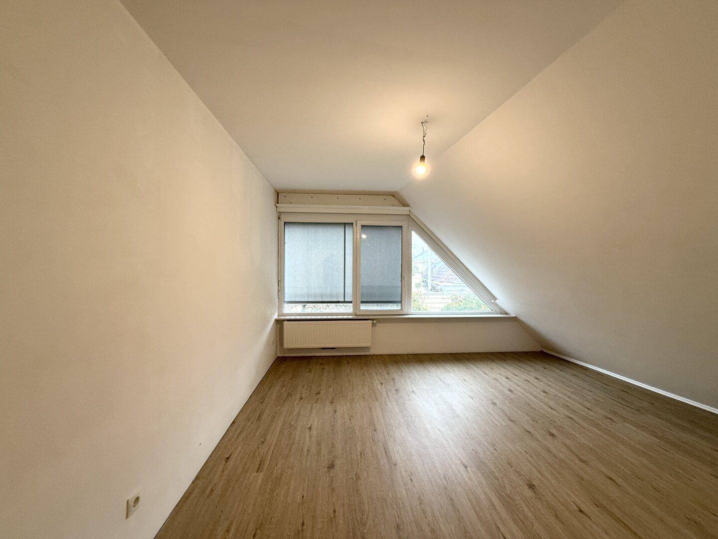 Dachgeschosszimmer mit schrägen Wänden, Laminatboden und einem großen Fenster für viel Tageslicht.