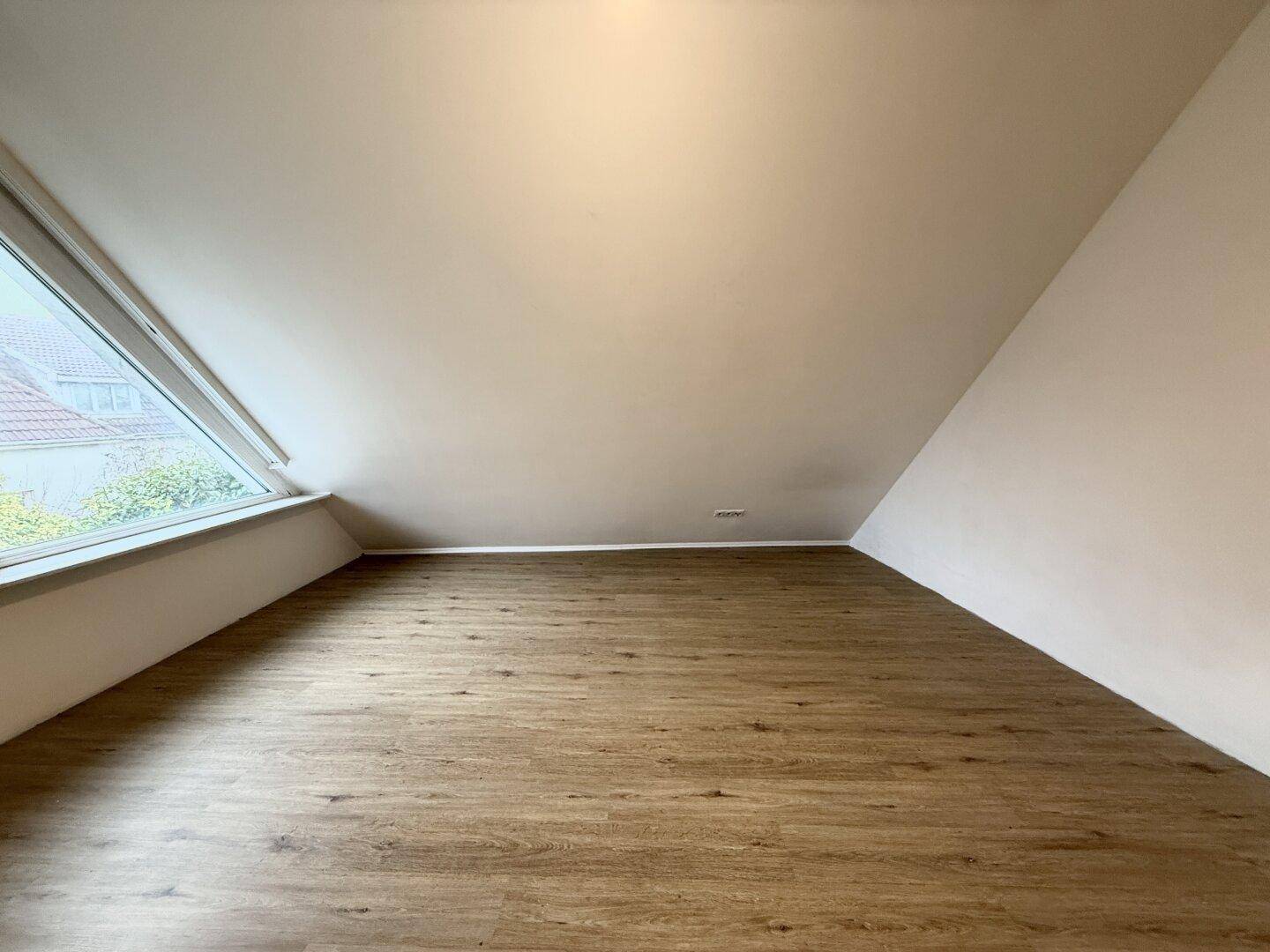 Dachgeschosszimmer mit schrägen Wänden und Laminatboden, das durch ein großes Fenster hell erleuchtet wird.