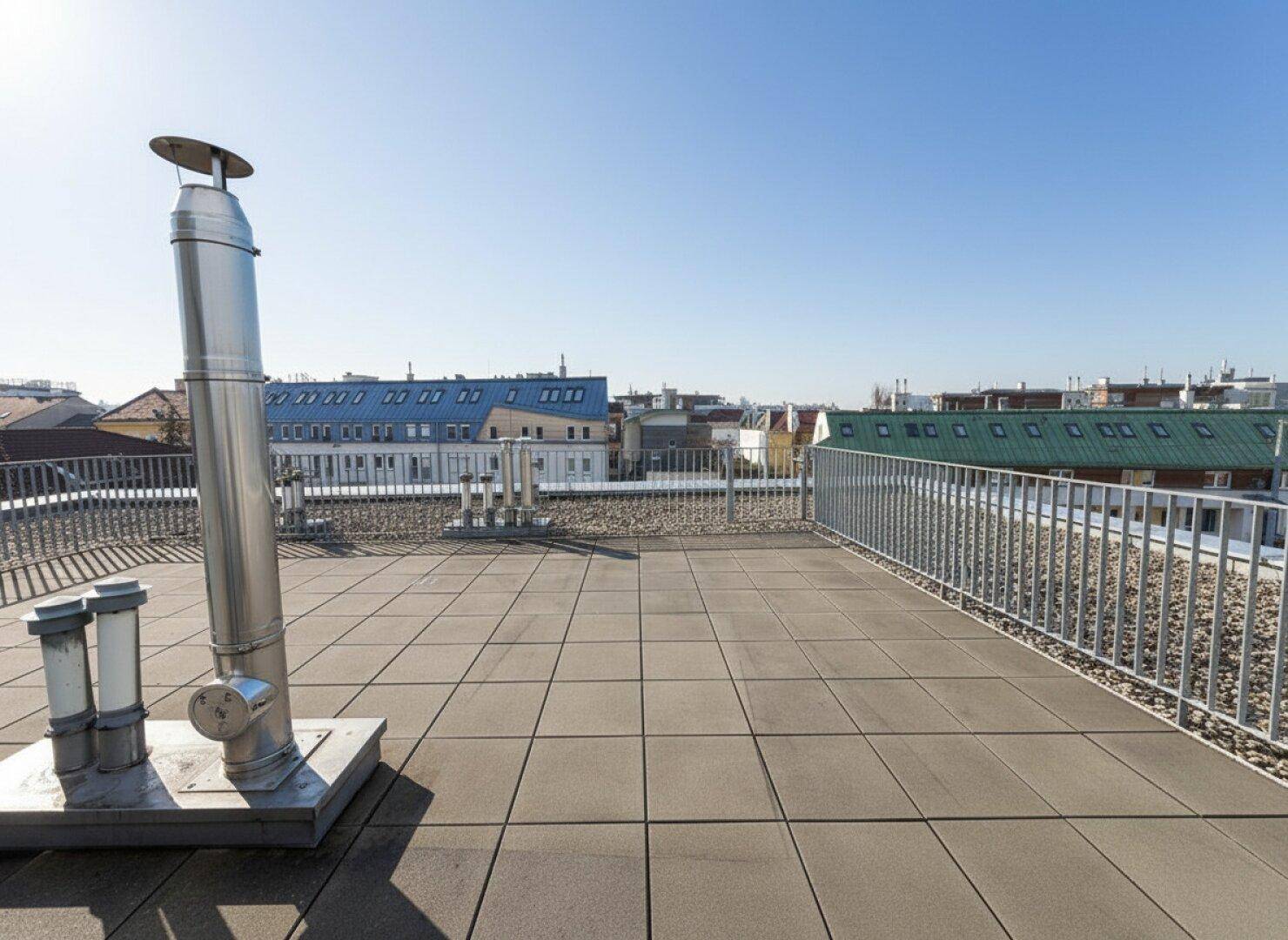 Weitläufige Dachterrasse mit Fliesenboden und Blick auf die umliegenden Gebäude unter blauem Himmel.