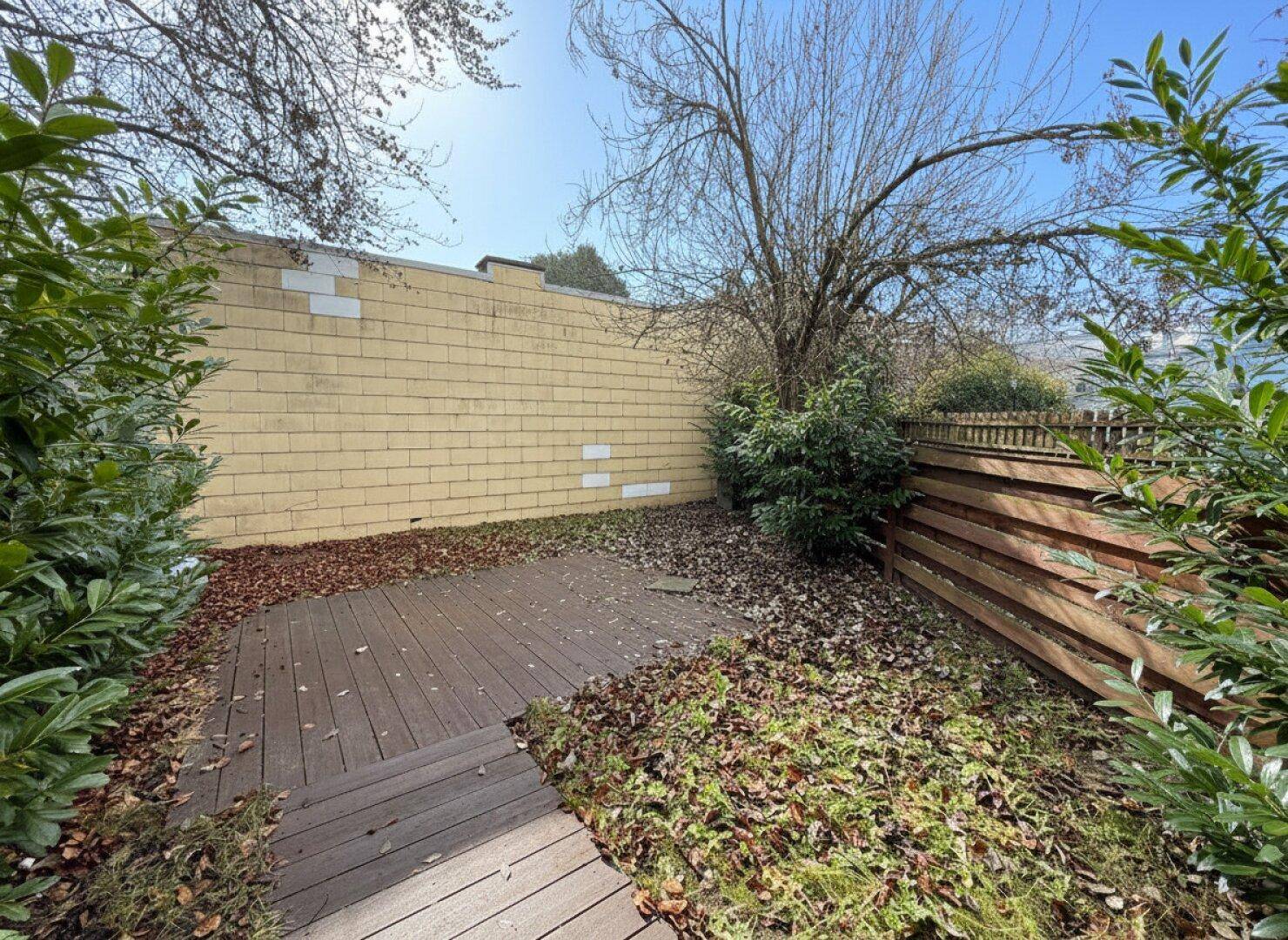 Holzterrasse im Garten, umgeben von Laub und grünen Pflanzen, mit einer gelben Wand und Holzzaun.