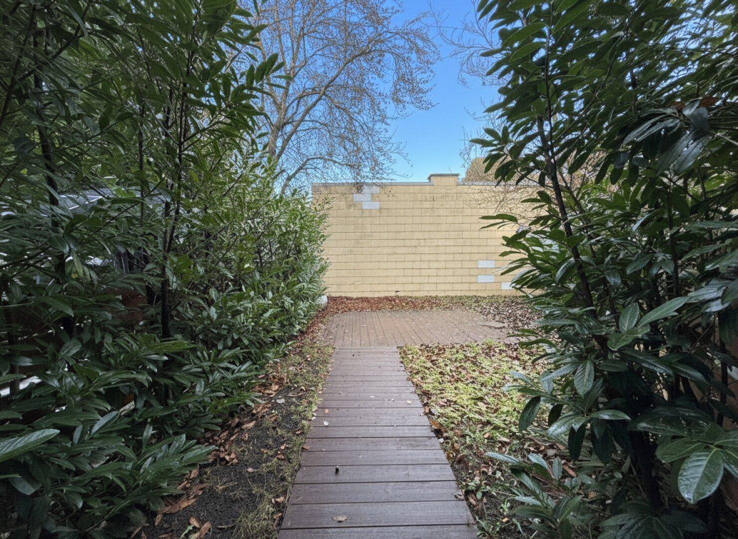 Holzweg führt durch grüne Sträucher zu einem privaten Gartenbereich mit gelber Mauer.