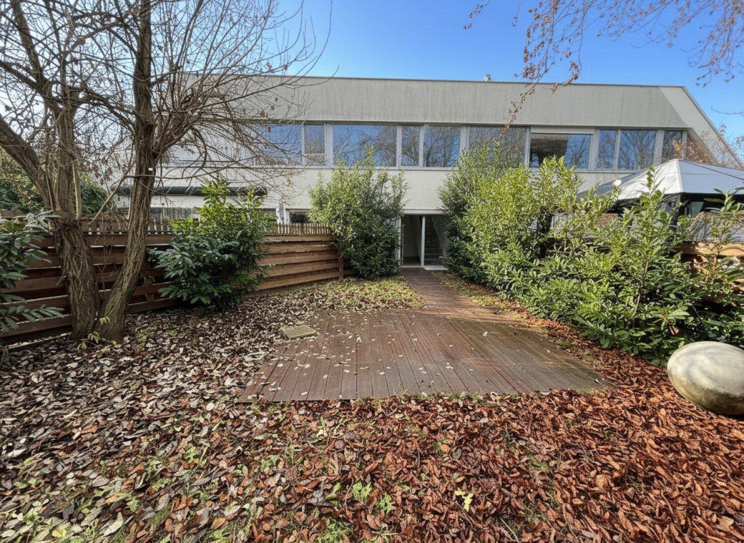 Gartenbereich mit Holzterrasse und Laub, im Hintergrund die moderne Fassade des Gebäudes.