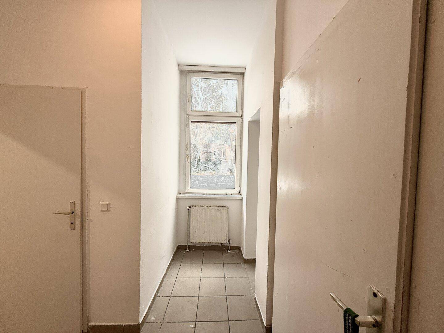 Schmaler Flur mit Fliesenboden und einem Fenster, das Tageslicht hereinlässt.
