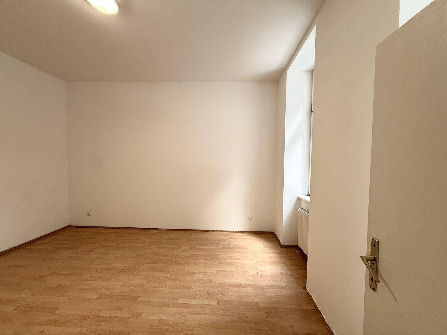 Leeres Zimmer mit Laminatboden und einem Fenster, das viel Tageslicht hereinlässt.