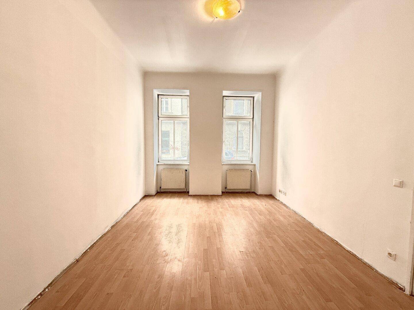 Helles, leeres Zimmer mit Laminatboden und zwei Fenstern, bereit zur individuellen Gestaltung.