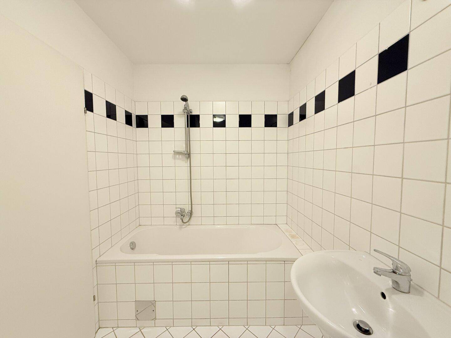 Badezimmer mit Badewanne und Duschfunktion, umgeben von weißen Fliesen und einem Waschbecken.