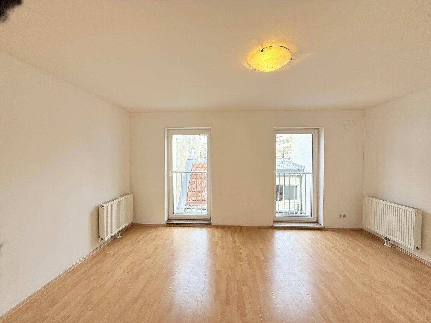 Helles Wohnzimmer mit Parkettboden und zwei französischen Balkonen, die viel Tageslicht hereinlassen.