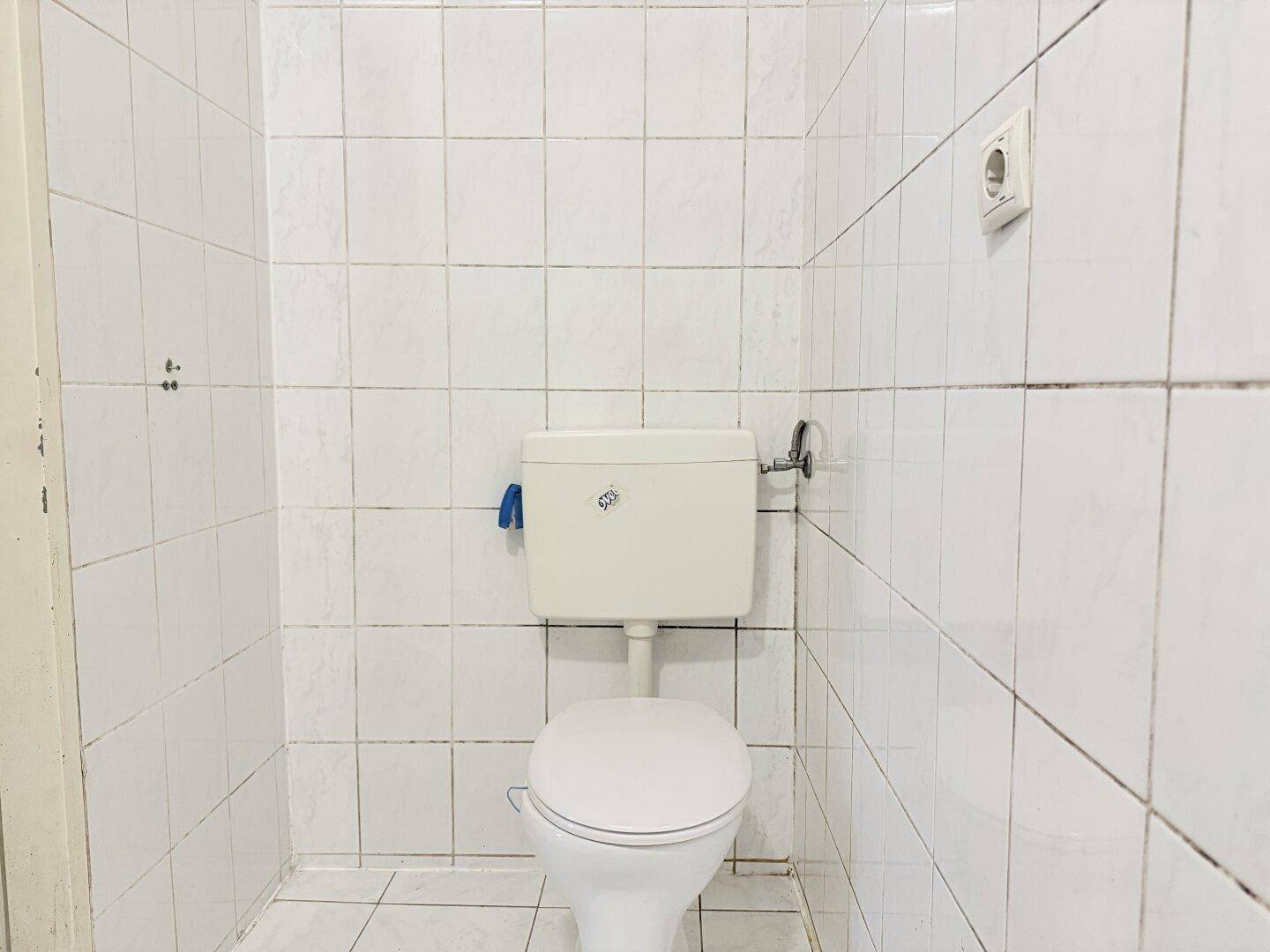 Separate Toilette mit weißen Fliesen und Standard-WC, in renovierungsbedürftigem Zustand.