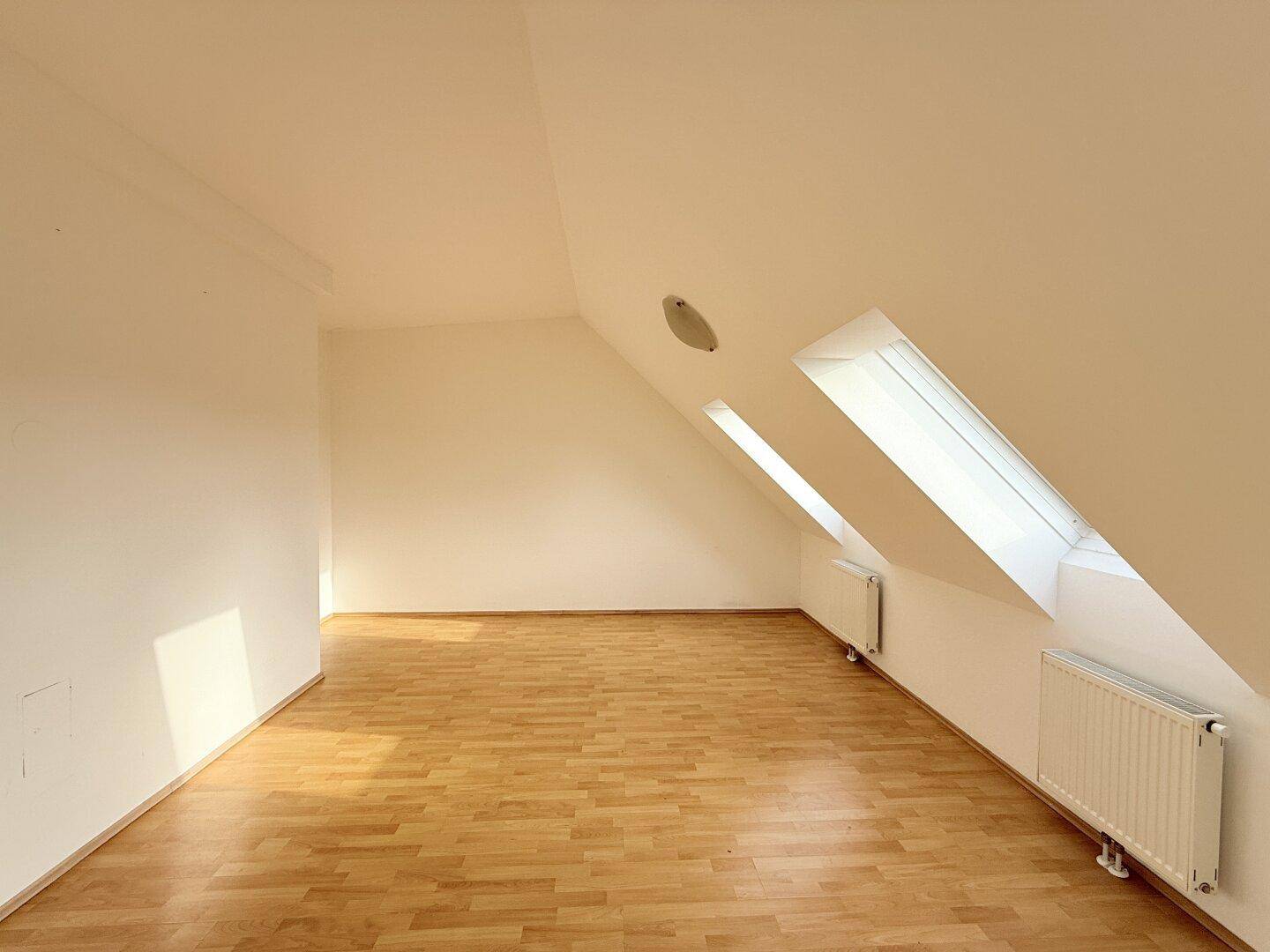 Leerer Raum im Dachgeschoss mit Parkettboden, Dachfenstern und Heizkörpern.