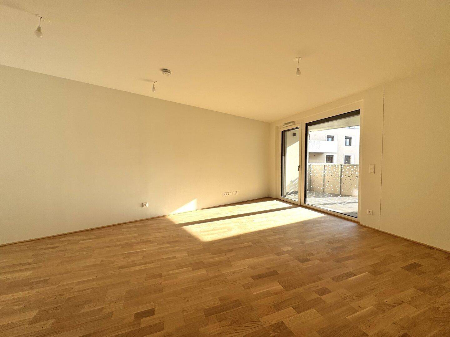 Lichtdurchfluteter, leerer Wohnbereich mit hellem Parkettboden und Zugang zum Balkon.