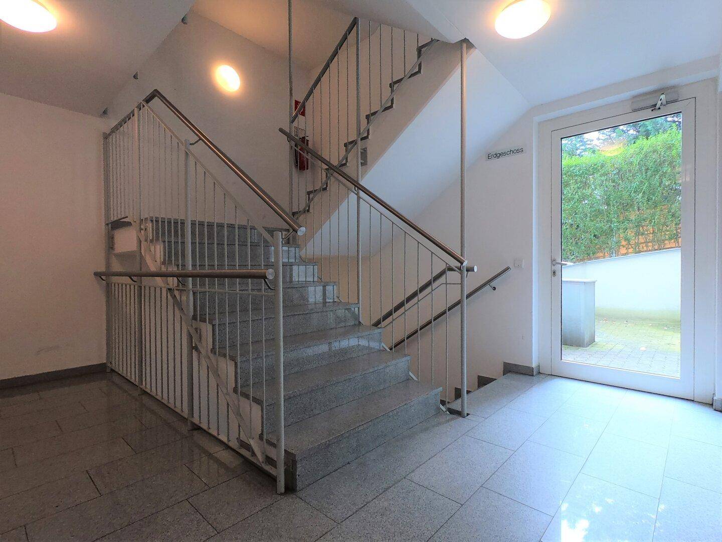 Helles Treppenhaus mit Granitfliesen und Metallgeländer, führt zum Erdgeschossausgang.