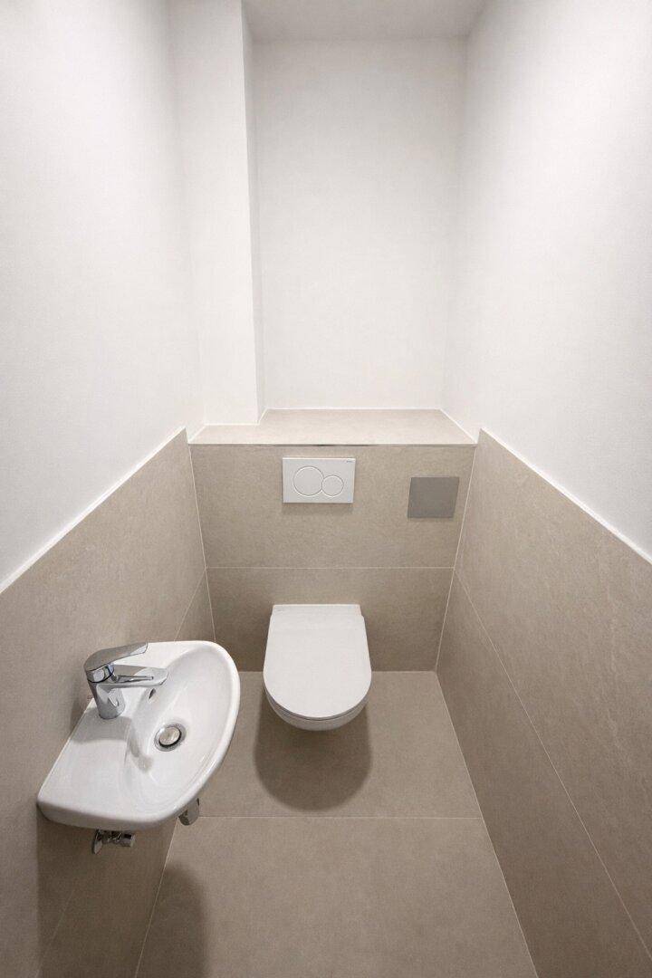 Kompaktes Gäste-WC mit Wand-WC und kleinem Waschbecken, halbhoch gefliest in Beige.