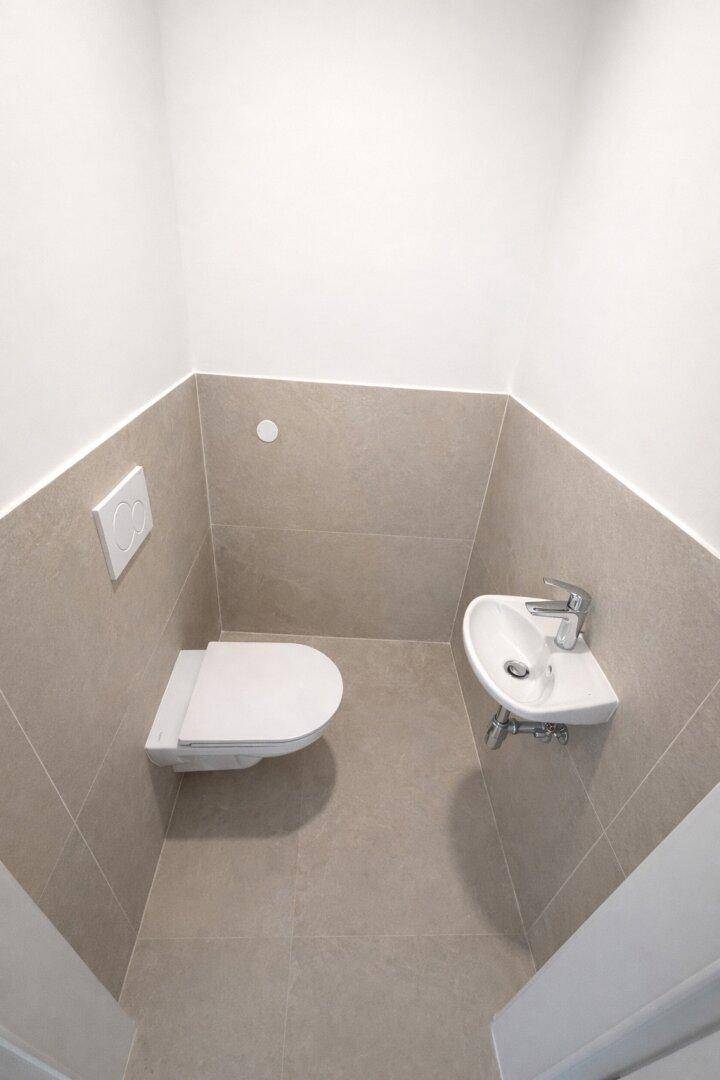 Kleines Gäste-WC mit beigefarbenen Fliesen, wandmontierter Toilette und kleinem Waschbecken.