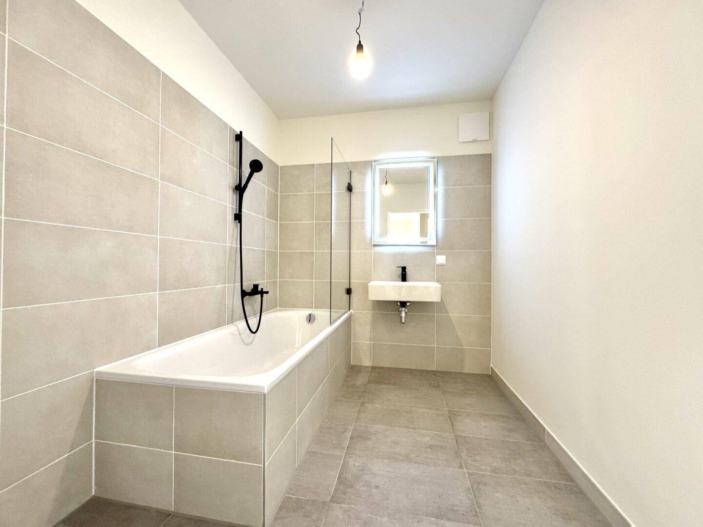 Modernes Badezimmer mit Badewanne, Duschwand und hellgrauen Fliesen.
