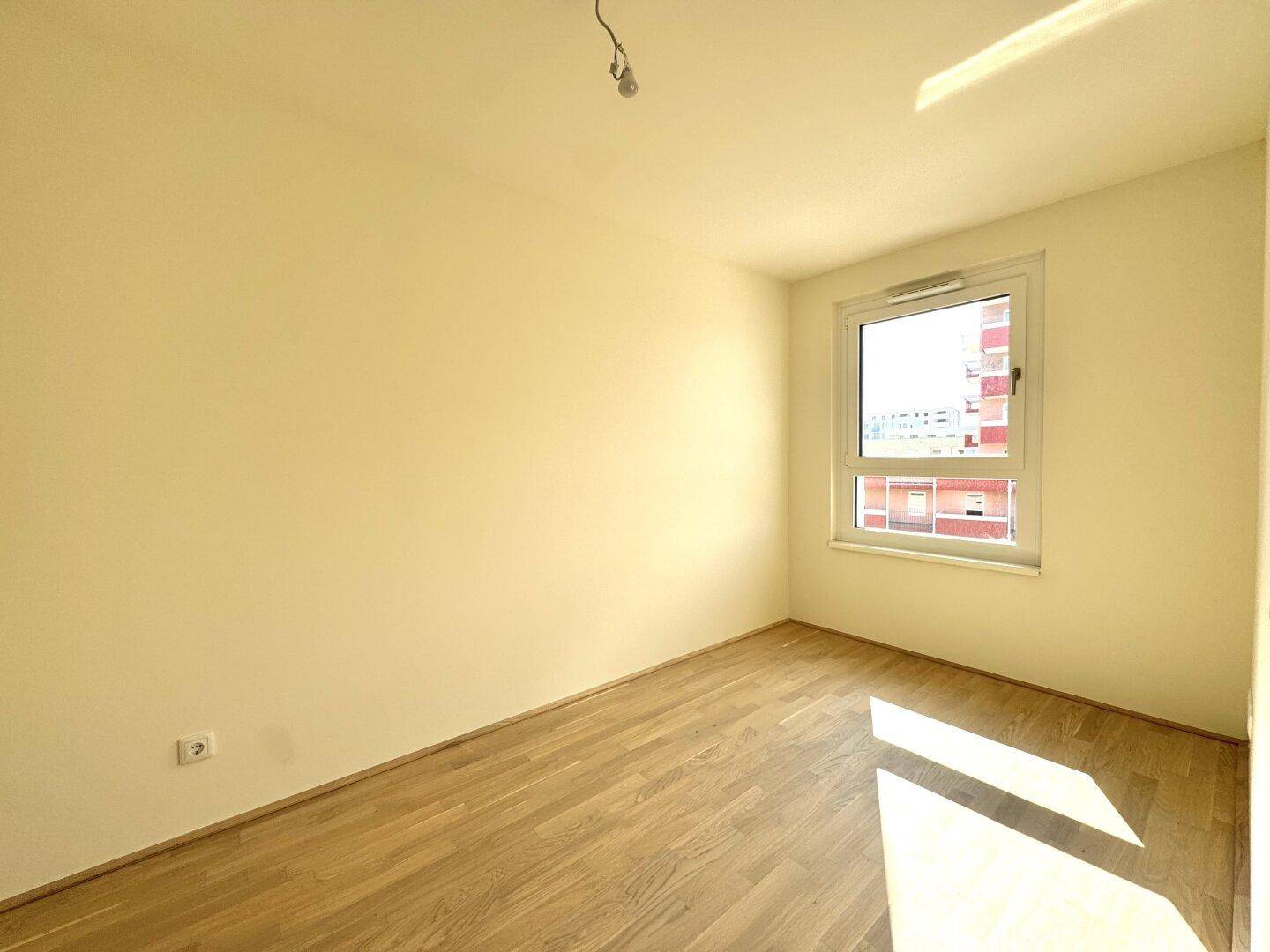 Leeres Schlafzimmer mit Parkettboden und großem Fenster, das viel Tageslicht hereinlässt.