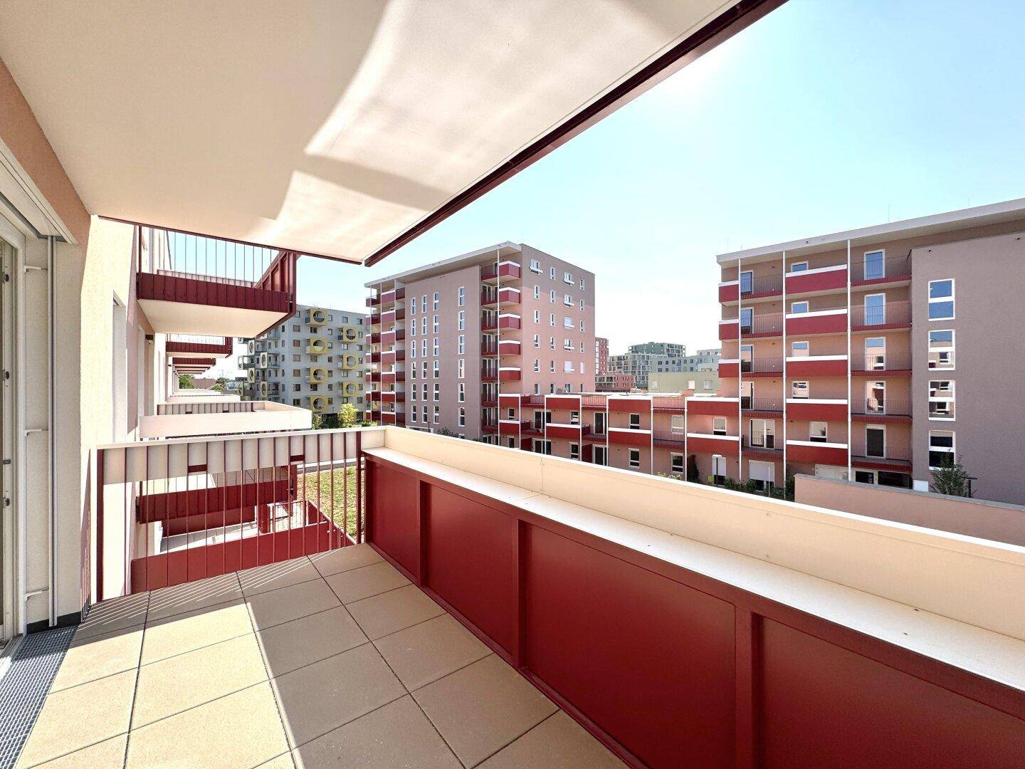 Geräumiger Balkon mit Fliesenboden und rotem Geländer, umgeben von modernen Gebäuden.