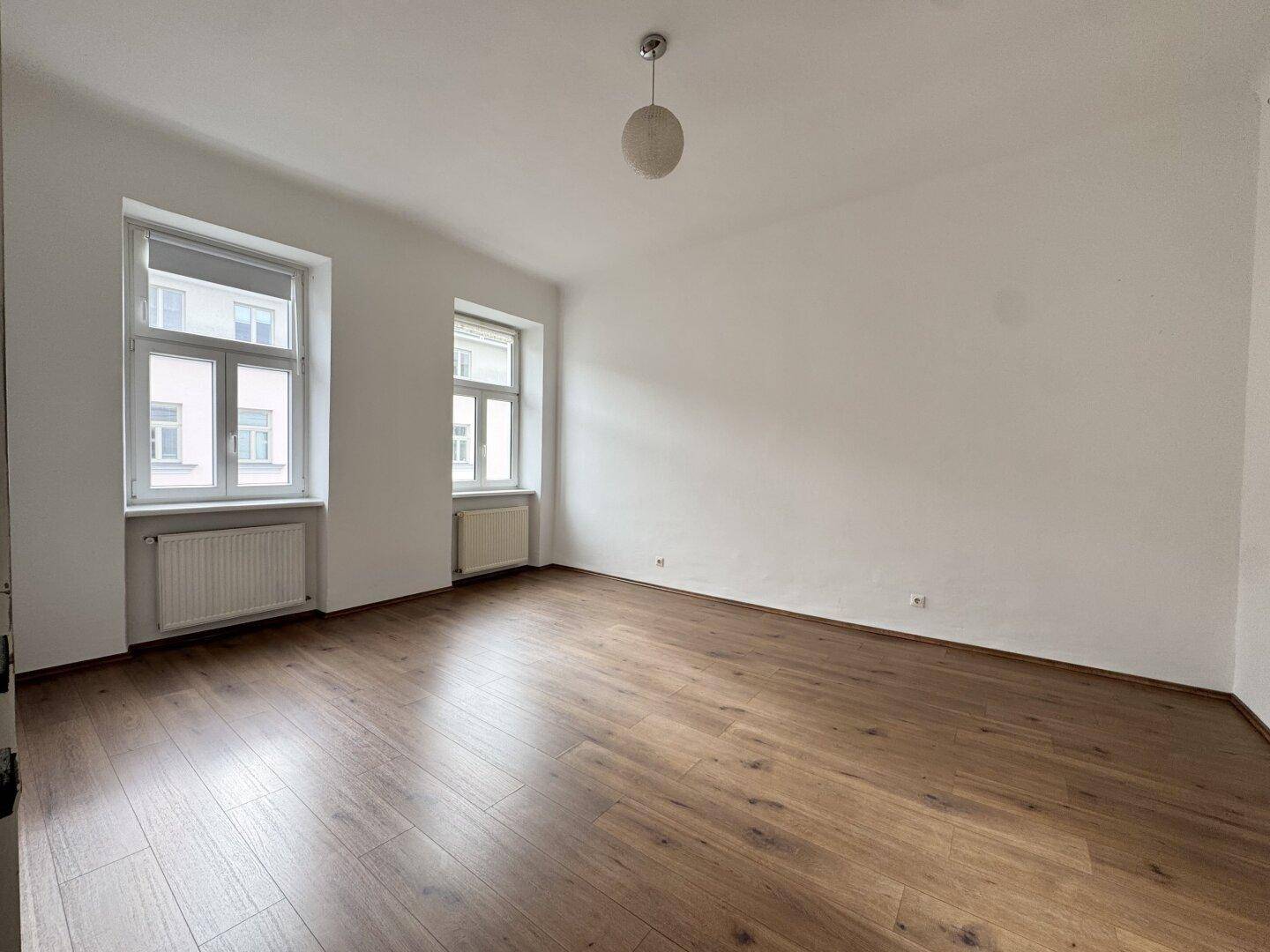 Geräumiger, heller Raum mit Holzoptik-Laminatboden und großen Fenstern, ideal für Wohnzwecke.