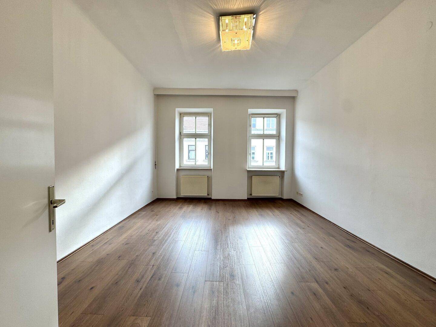 Heller, leerer Raum mit Laminatboden und zwei Fenstern, bereit zur individuellen Gestaltung.