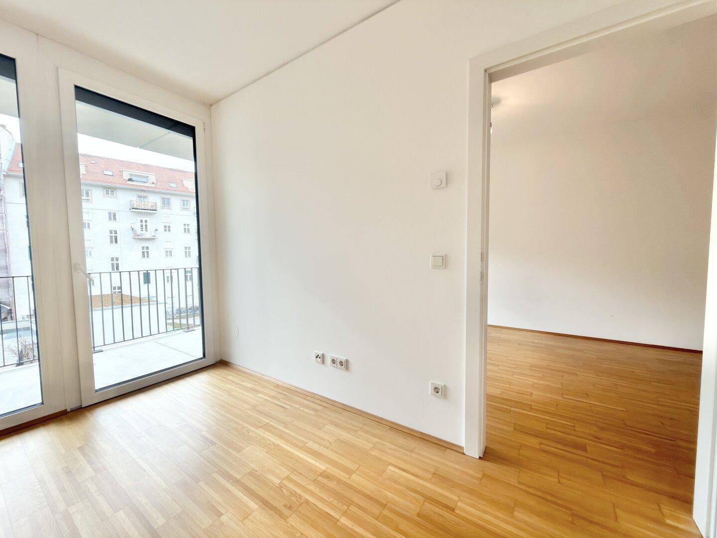 Heller Raum mit Parkettboden und großen Fenstern, die zu einem französischen Balkon führen.