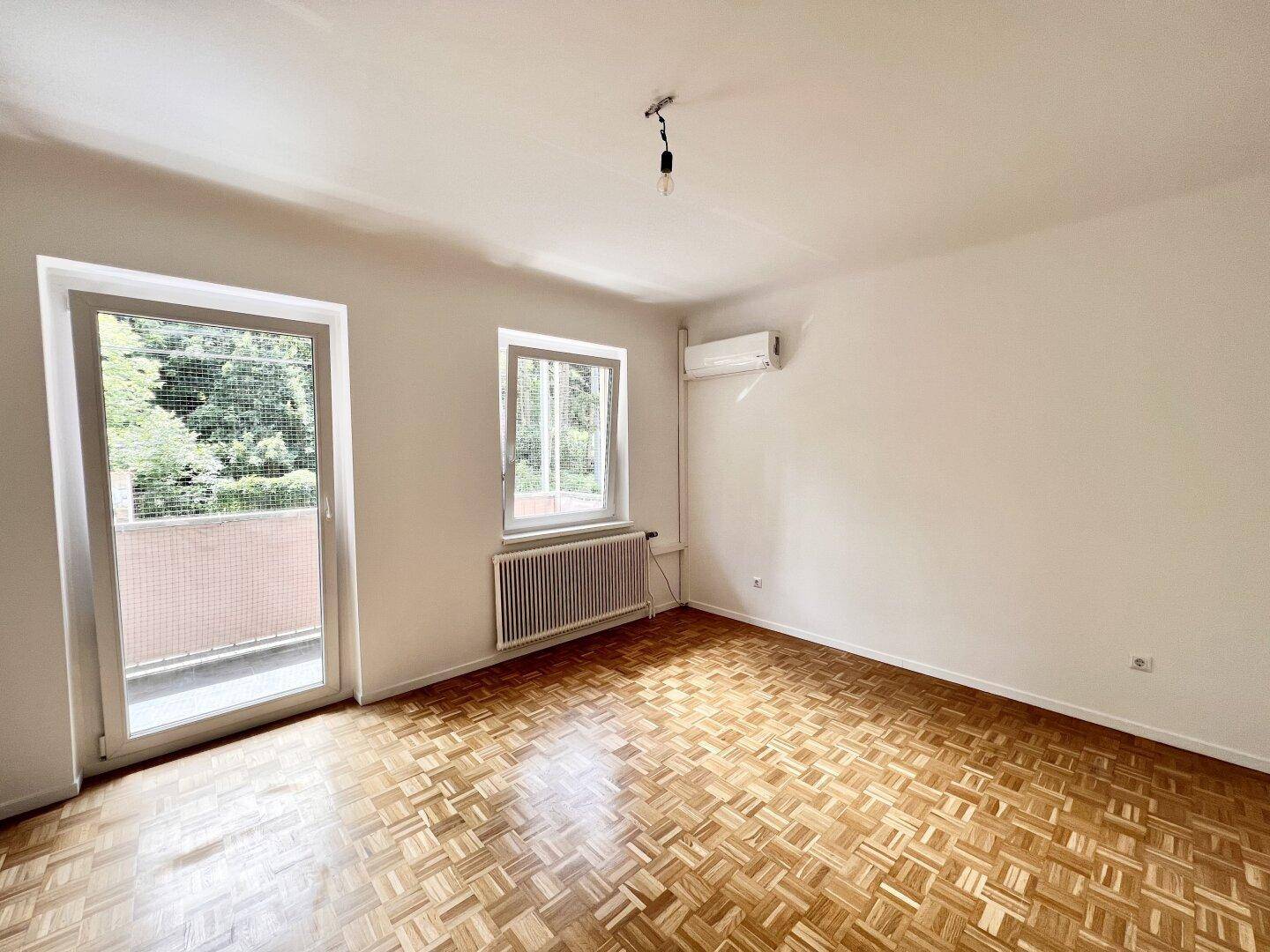 Leeres Schlafzimmer mit Parkettboden, Fenster und Zugang zum Balkon, bereit zur Einrichtung.