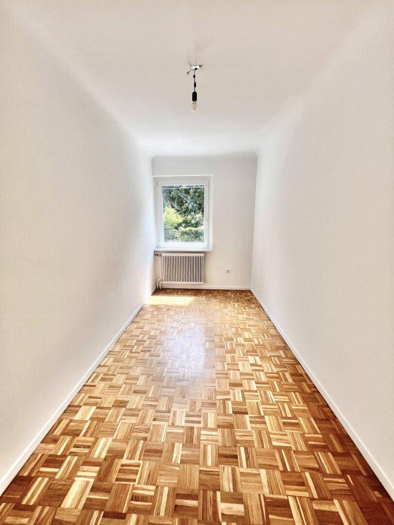 Leeres Zimmer mit Parkettboden und großem Fenster, ideal als Kinder- oder Arbeitszimmer.