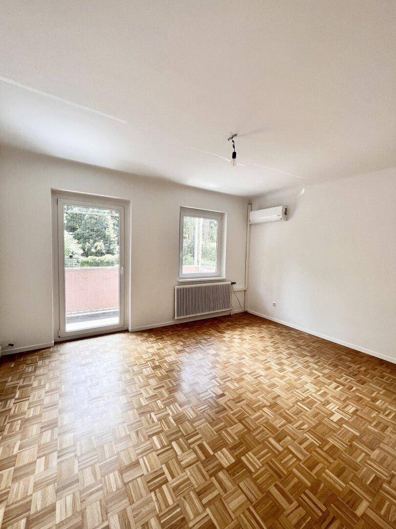 Leeres Schlafzimmer mit Parkettboden, Fenster und Zugang zum Balkon, bereit zur Einrichtung.