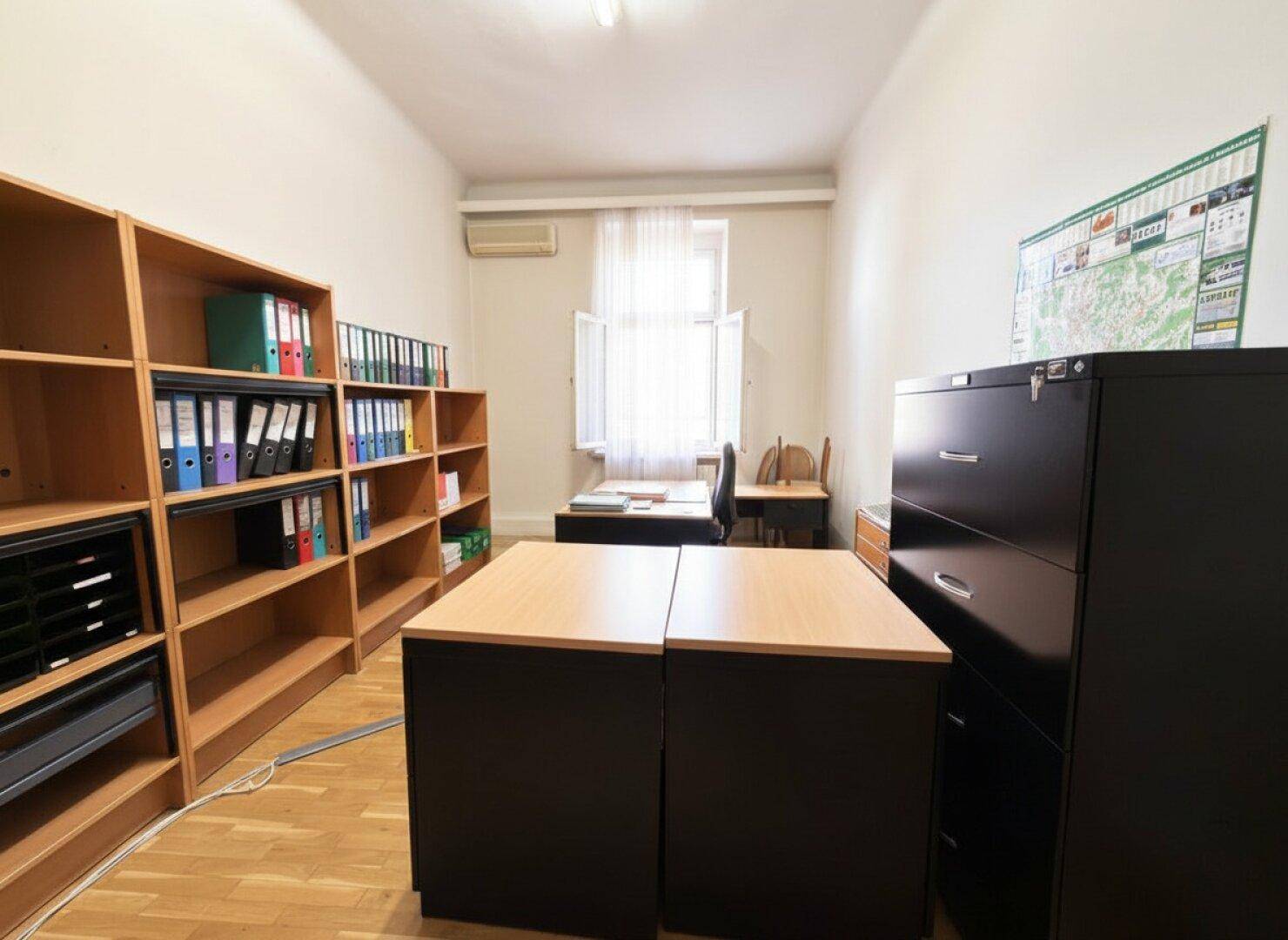 Funktionales Büro mit Regalen, Aktenschränken und einem kleinen Arbeitsbereich am Fenster.