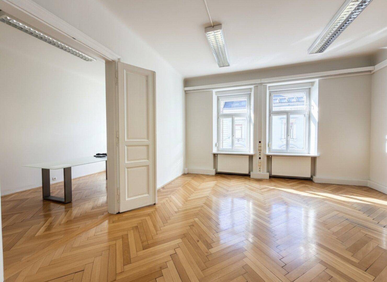 Helles, leeres Büro mit Fischgrätparkett und großen Fenstern, bereit zur Einrichtung.