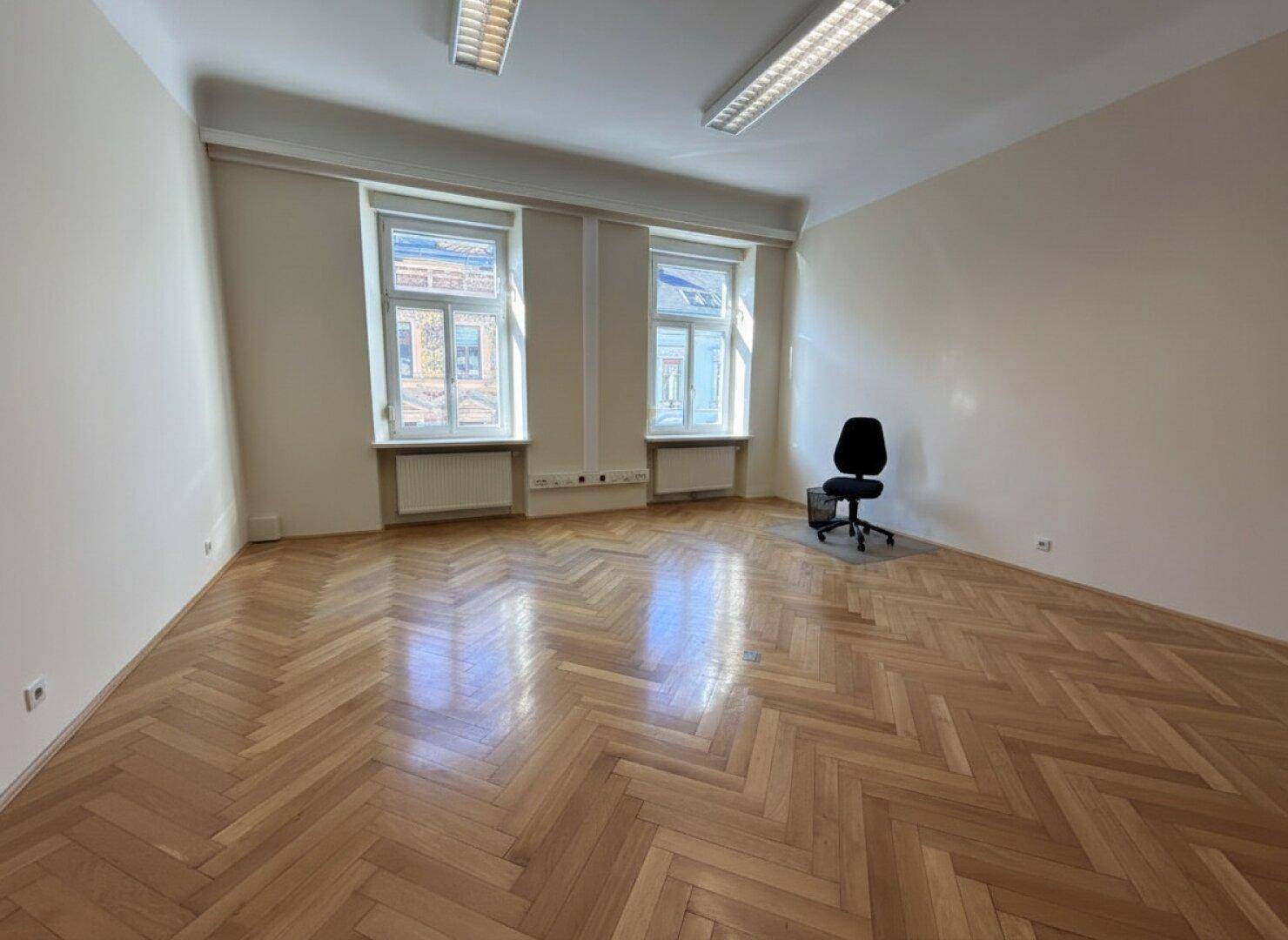Leeres Büro mit glänzendem Fischgrätparkett und zwei großen Fenstern für viel Tageslicht.