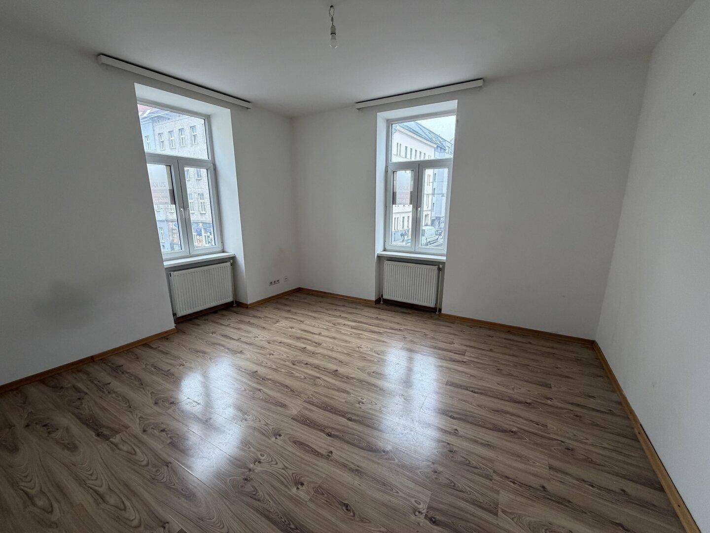 Heller, leerer Raum mit Laminatboden und zwei großen Fenstern zur Straße.