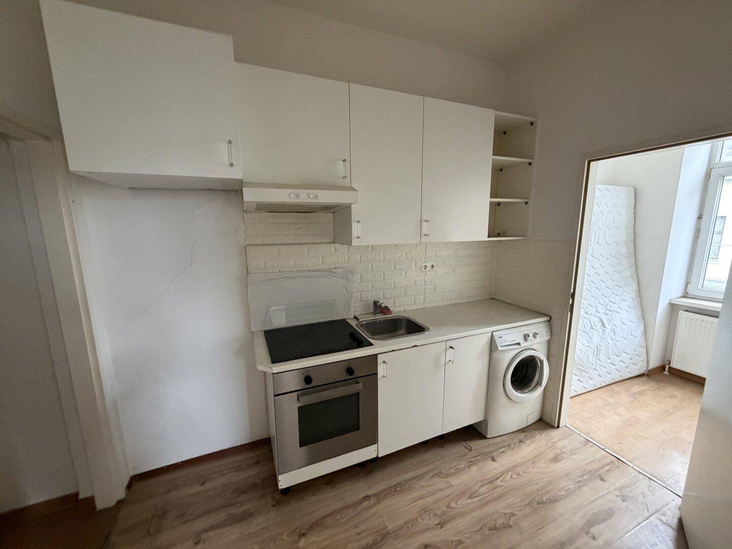 Kleine Einbauküche mit weißen Schränken, Kochfeld, Backofen und integrierter Waschmaschine.