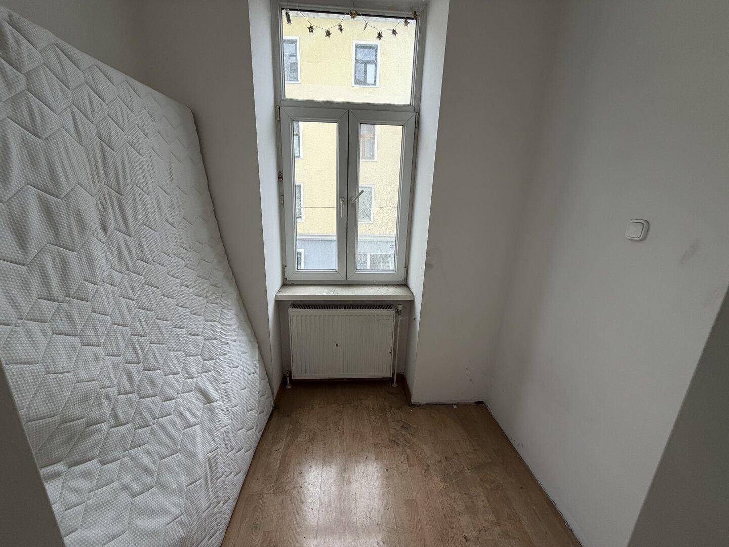 Kleines Schlafzimmer mit Fenster, Heizkörper und einer Matratze an der Wand.