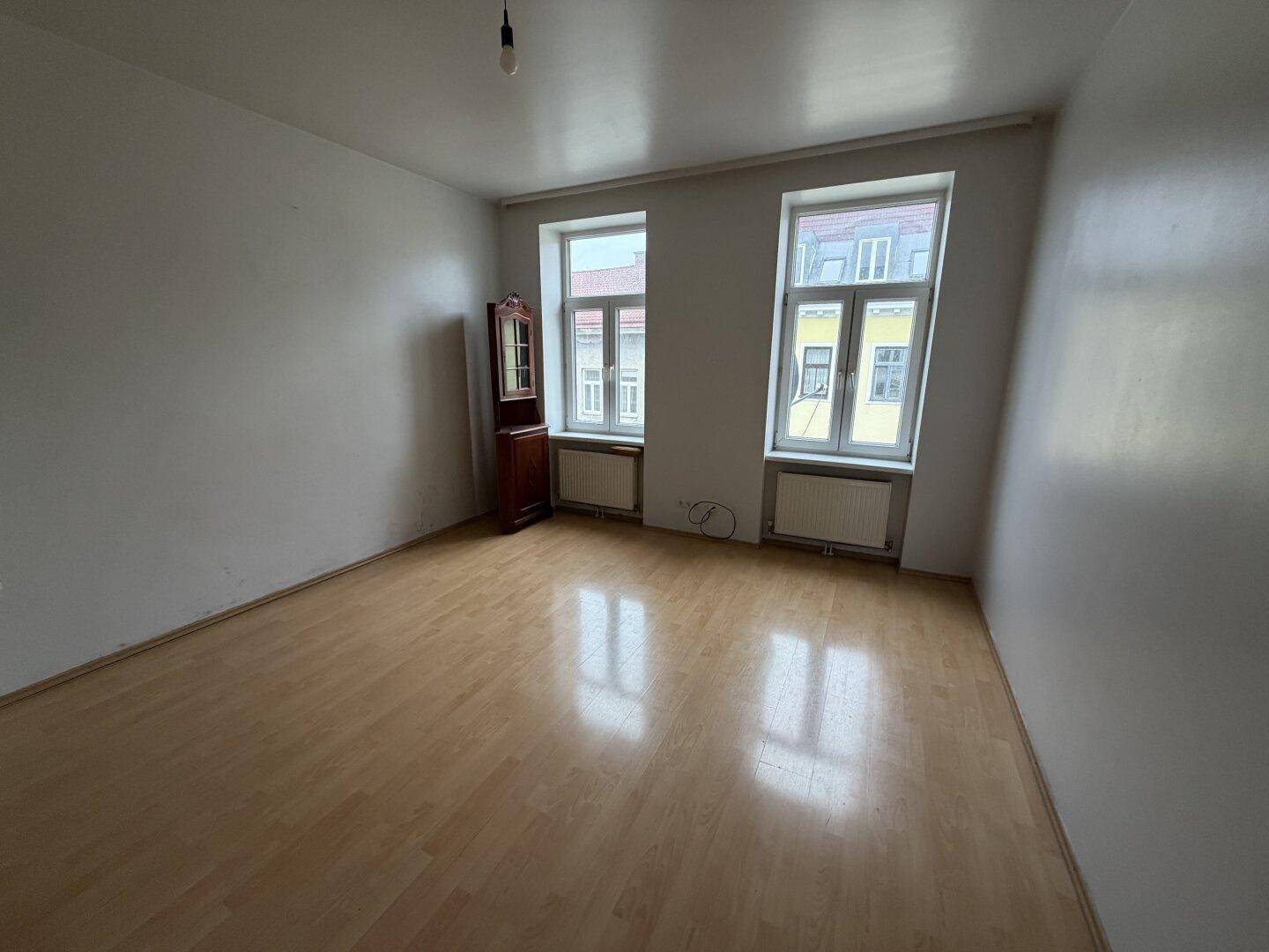 Helles, leeres Zimmer mit Laminatboden und zwei Fenstern, bereit zur individuellen Gestaltung.