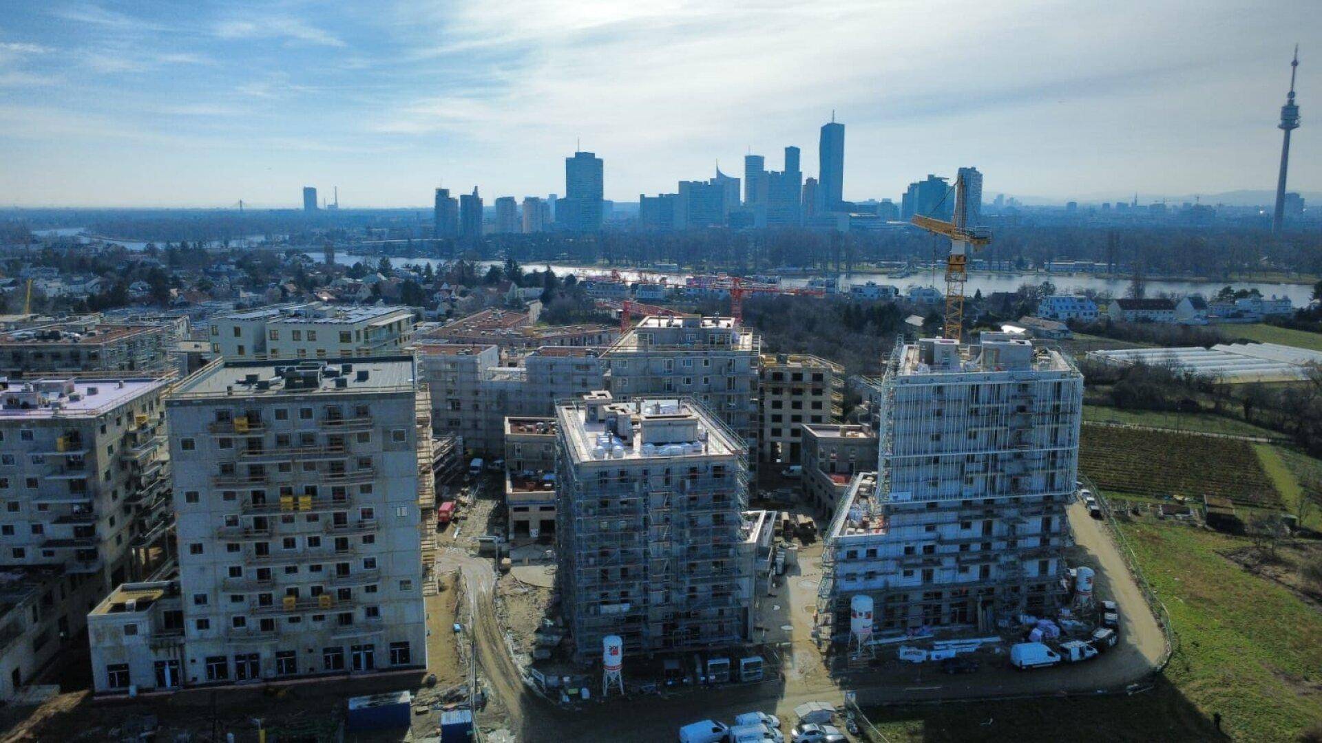 Übersicht der Baustelle mit unfertigen Gebäuden und der Donau sowie der Stadt im Hintergrund.