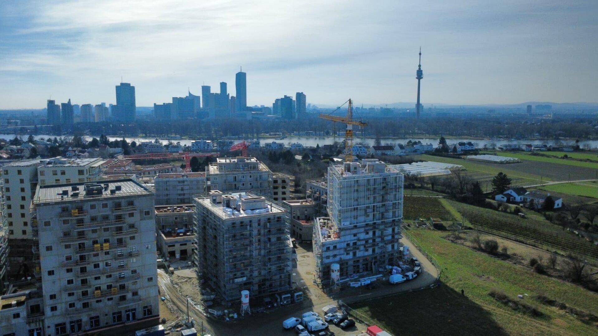 Weitläufige Drohnenansicht einer Wohnbaustelle mit Kränen, Feldern und der beeindruckenden Skyline einer Großstadt am Horizont.