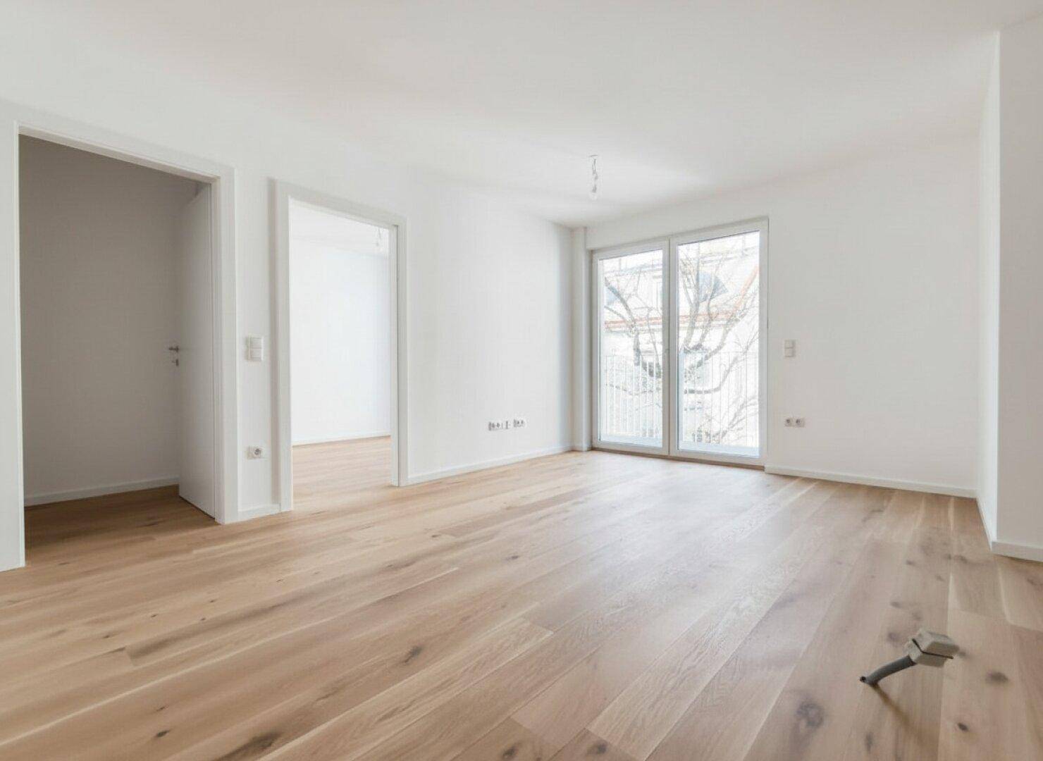 Helles, leeres Zimmer mit neuem Holzparkett und Zugang zu einem Balkon.