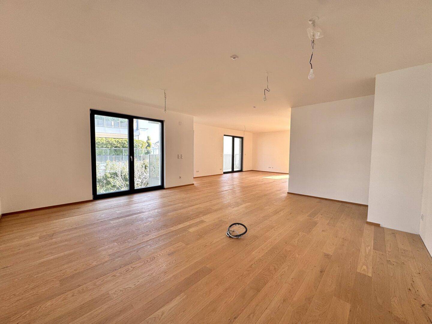 Geräumiges, leeres Wohnzimmer mit hellem Holzboden und großen Fenstertüren für viel Tageslicht.
