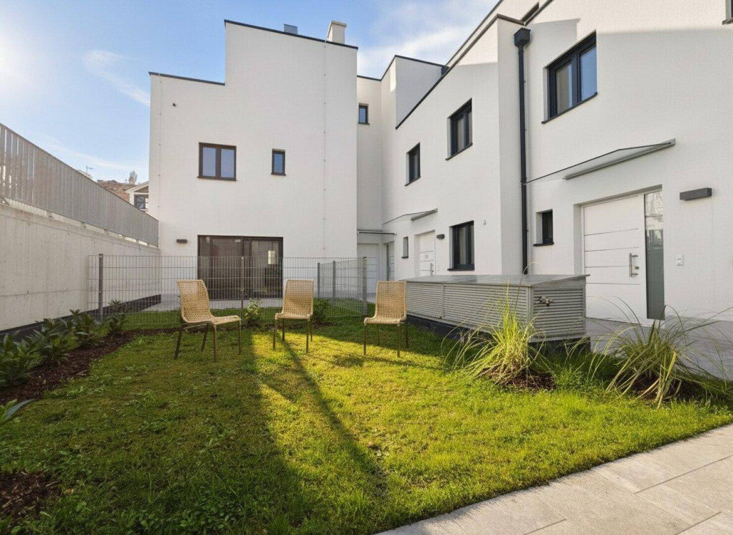 Moderner Neubau mit gepflegtem Rasen und Sitzgelegenheiten im Gartenbereich unter blauem Himmel.