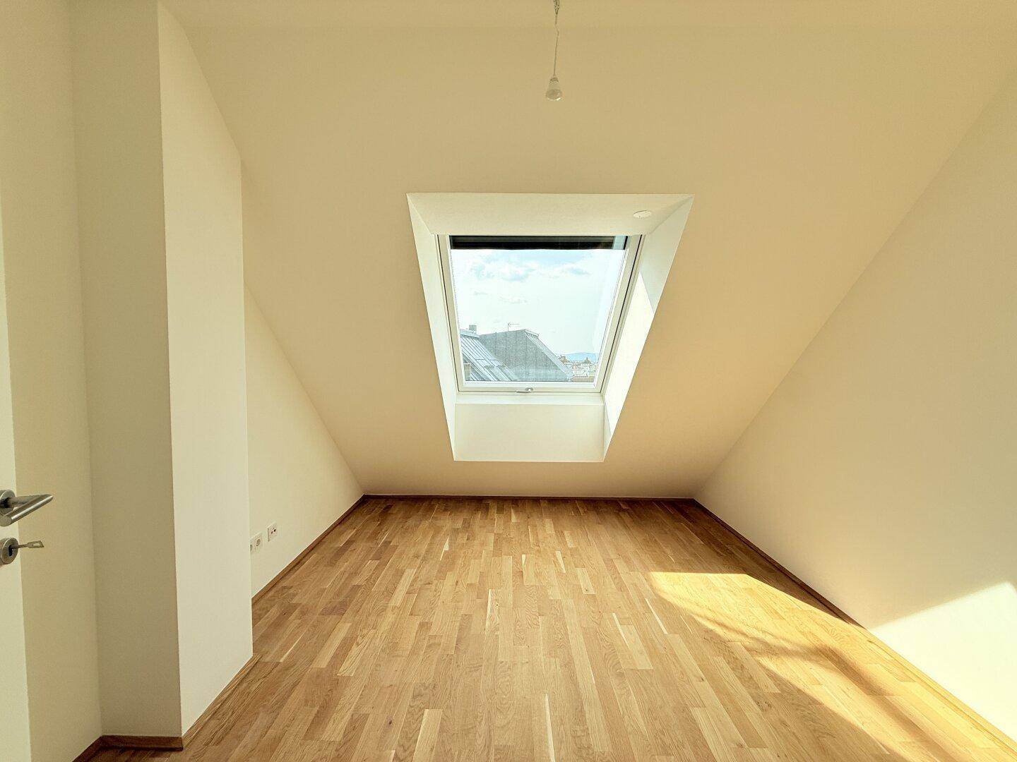 Helles Schlafzimmer im Dachgeschoss mit einem großen Dachfenster und Parkettboden, sonnig.
