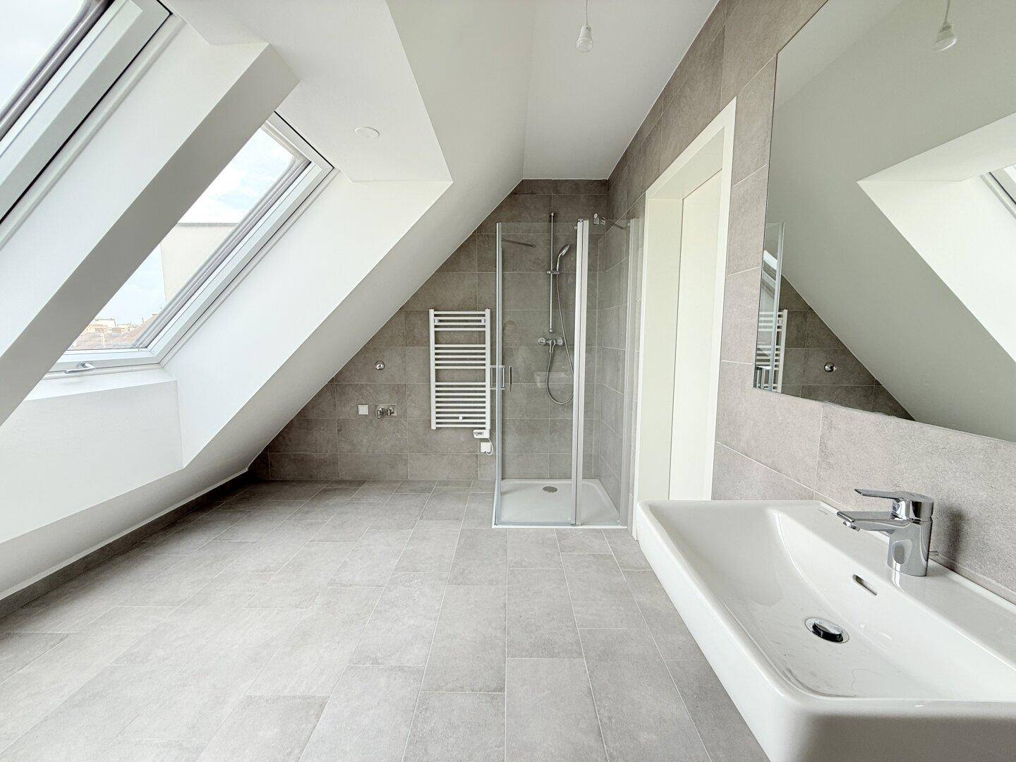 Modernes Badezimmer im Dachgeschoss mit Dusche, Waschbecken und Dachfenstern, hell gefliest.