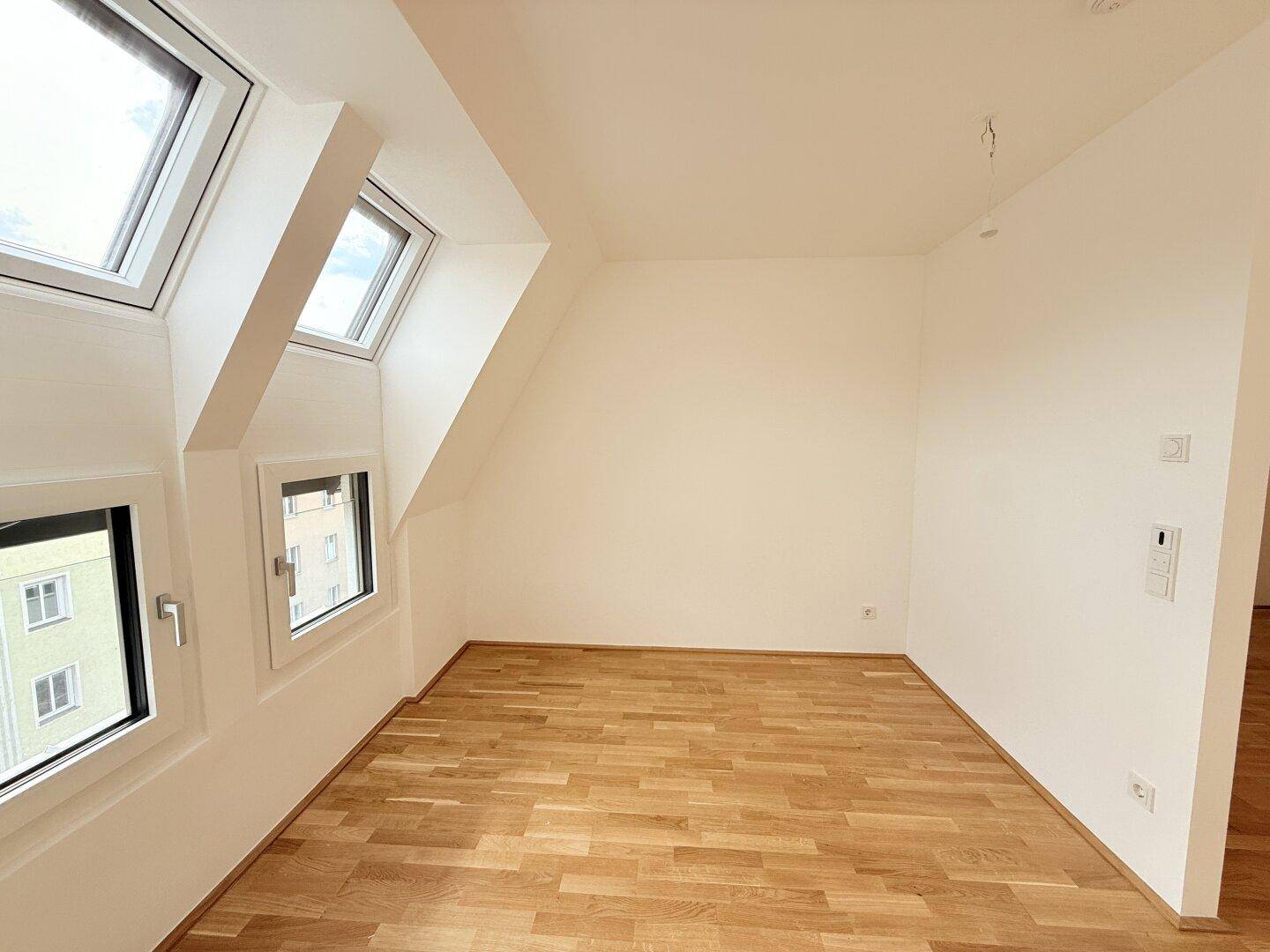 Helles Zimmer im Dachgeschoss mit Parkettboden, schrägen Wänden und mehreren Fenstern für viel Tageslicht.